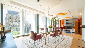 A spacious living area with a glass dining table, comfortable seating, and a large window offering a view of a historic building. - 4 Private Homes for rent to live the sparkling Remoise life. (Reims)