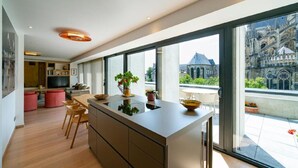 A modern kitchen with a large island, wooden flooring, and a view of a historic building through large windows. - 4 Private Homes for rent to live the sparkling Remoise life. (Reims)