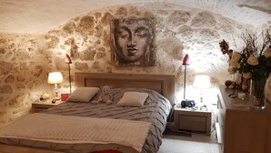 A cozy bedroom with a stone wall, a bed with a patterned bedspread, two bedside lamps, a nightstand with a lamp, a vase with flowers, and a teddy bear on the bed. - Maison de Village Atypique (Vidauban)