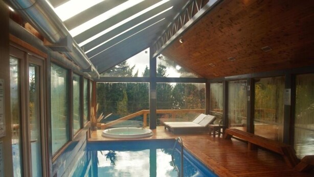 A modern indoor pool with glass walls, wooden deck, and a hot tub.