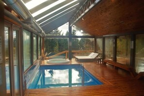Ein moderner, überdachter Pool mit Glaswänden, Holzterrasse und Whirlpool.