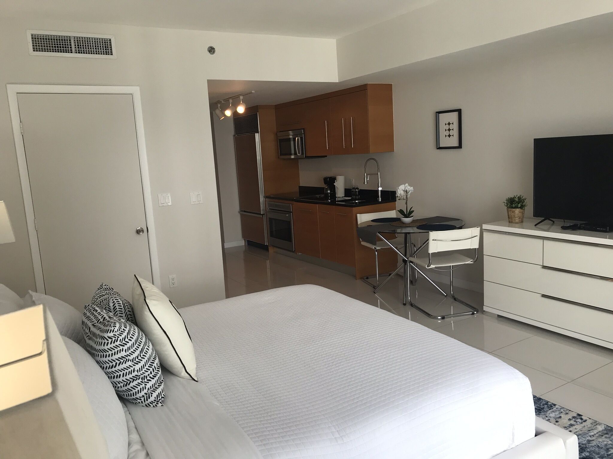 A modern hotel room with a large bed, a small kitchenette, a dining area, and a TV.