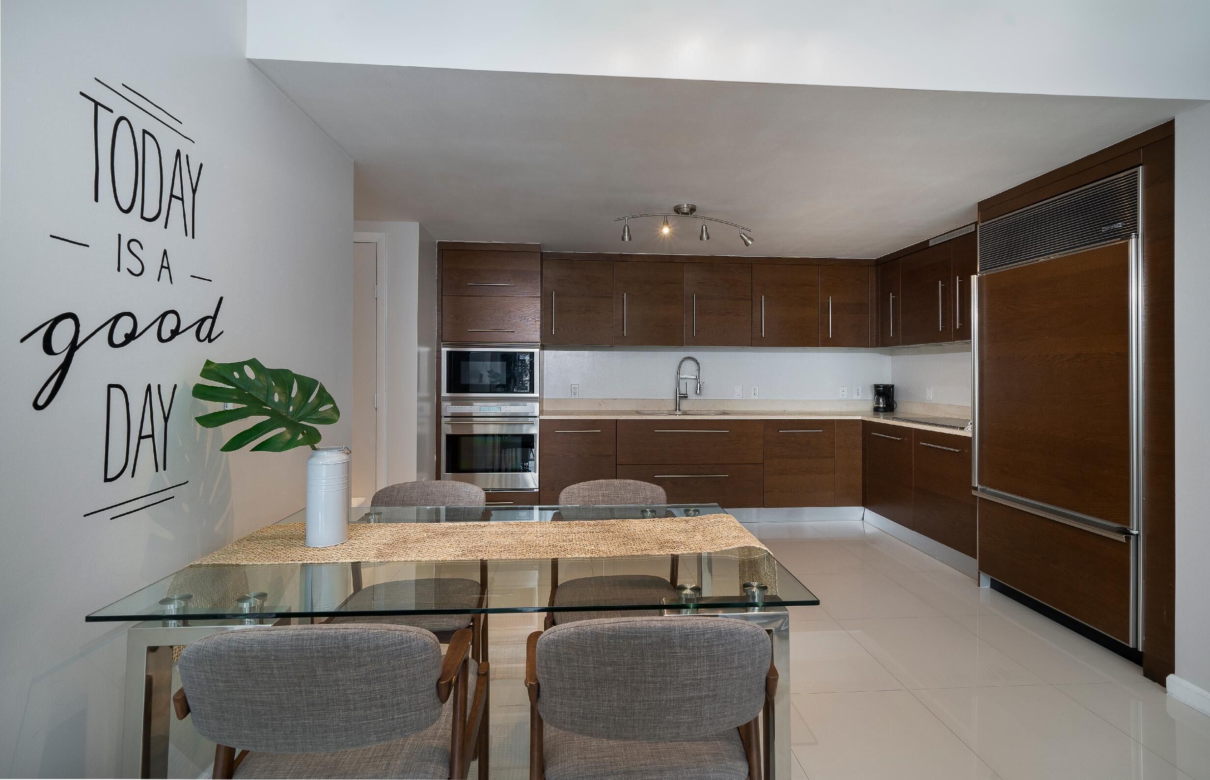 A modern kitchen with a glass dining table, wooden cabinets, and a stainless steel refrigerator.