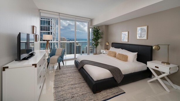 Ein modernes Schlafzimmer mit einem großen Bett, einem Fernseher, einem Schreibtisch und einem Balkon mit Blick auf die Stadt.