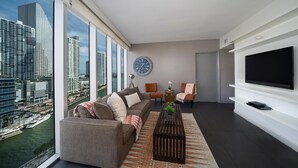 A modern living room with a large window, a sofa, two chairs, a coffee table, and a flat-screen TV. - Presidential River & City View (Miami)