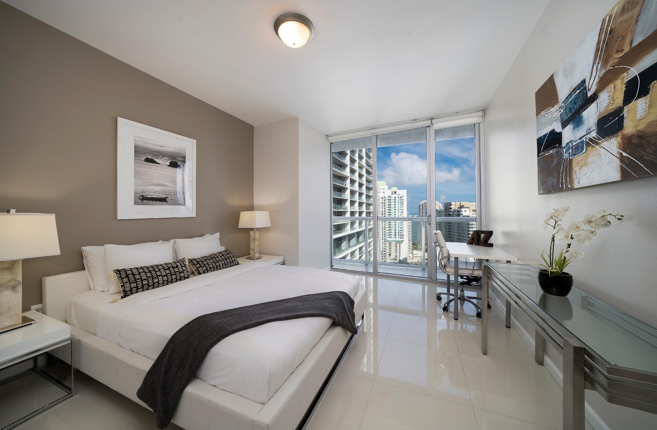 Una habitación de hotel moderna con una cama grande, un escritorio y vistas de la ciudad a través de ventanas de gran altura.