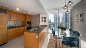 A modern kitchen with wooden cabinets, a granite countertop, and a built-in island with a sink. There is a dining area with a glass table and chairs, and a large window offering a city view. - Presidential River & City View (Miami)
