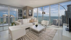 A modern living room with a large window, white furniture, and a view of the city and water. - Presidential Ocean View (Miami)