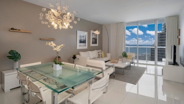 Ein modernes Wohnzimmer mit einem Glastisch, weißen Sofas, einem großen Fenster mit Stadtblick und einer dekorativen Chandelier.