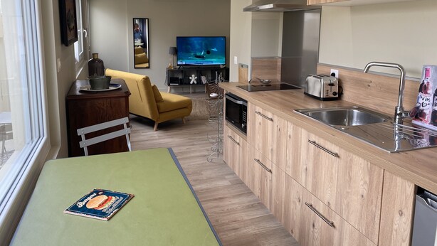 Een moderne keuken met houten kastjes, een inbouwoven en een spoelbak. Er is een eethoek met een gele stoel en een tafel. Een flatscreen televisie hangt aan de muur.