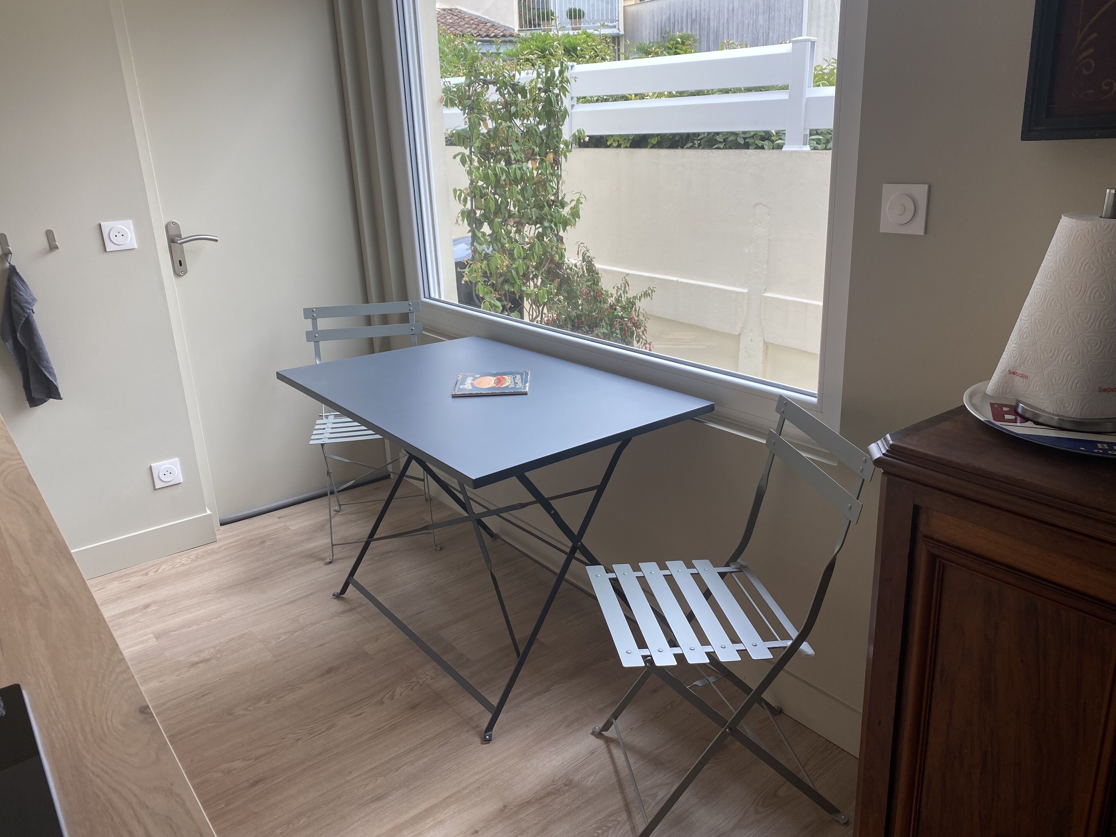 Un petit balcon avec une table et des chaises, une vue sur une clôture blanche et sur la verdure, ainsi qu’un meuble en bois.