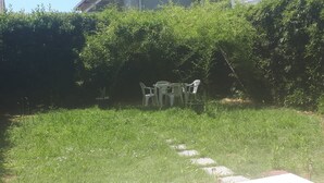 Un espace patio doté d’un sol carrelé, entouré d’une pelouse et d’une haie. On y trouve des chaises en plastique et une petite table.