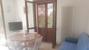 Una habitación con una mesa de comedor redonda, sillas blancas, un sofá azul con cojines, un televisor sobre un mueble bajo y una ventana con persianas.