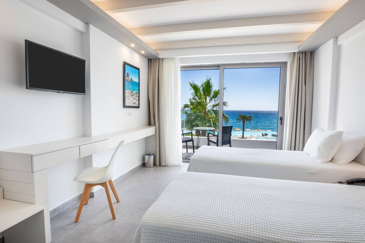 A hotel room with two beds, a desk, a chair, a TV, and a balcony with a view of the sea.