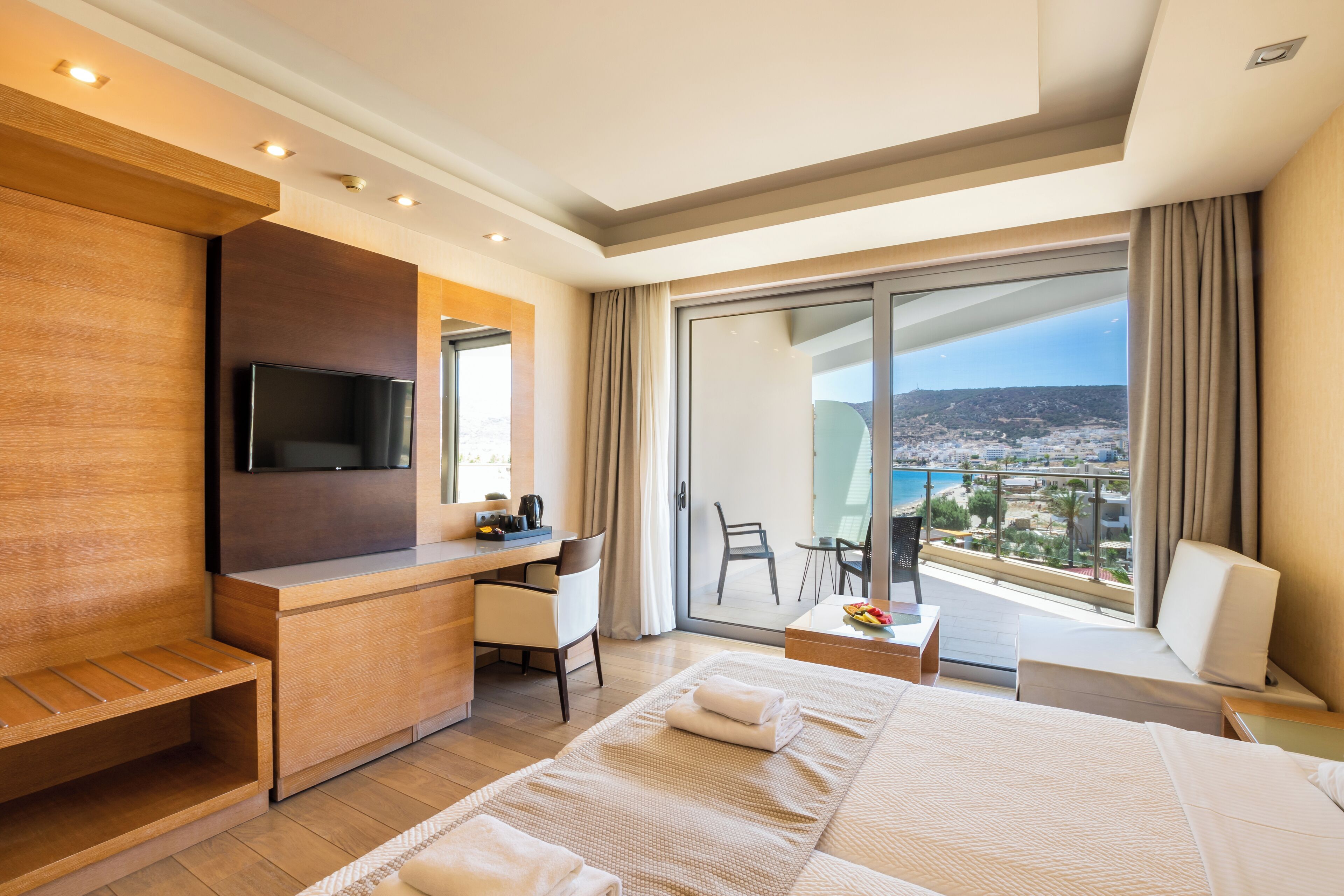 Ein modernes Hotelzimmer mit einem großen Bett, einem Schreibtisch mit Flachbildfernseher, einem Balkon mit Blick auf die Stadt und einer Sitzecke mit Tisch und Stühlen.
