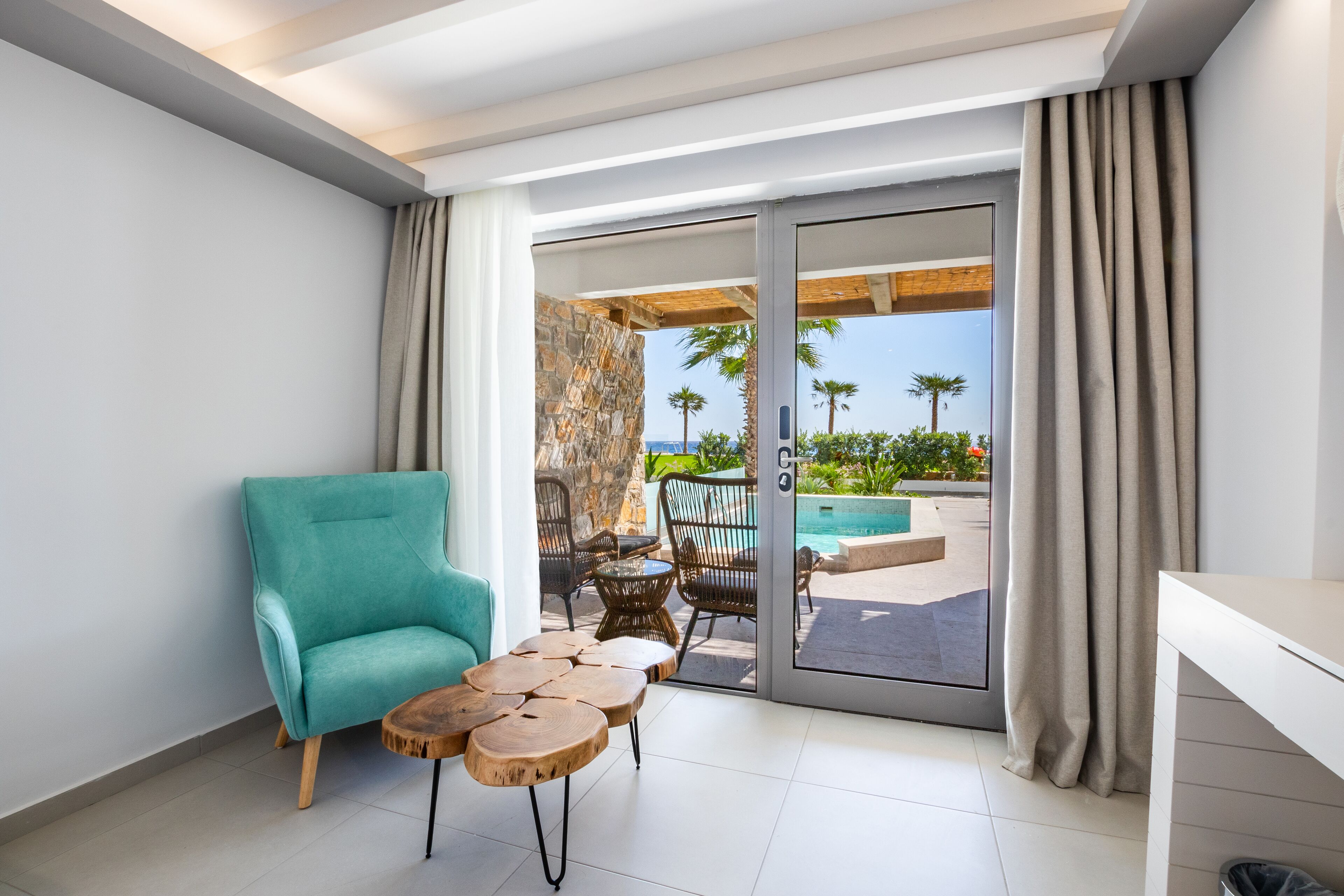 A modern room with a teal armchair, a small wooden table, and a glass door leading to an outdoor area with a pool and palm trees.