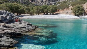 A rocky shoreline with clear turquoise water, a person sitting on the rocks, and a mountainous backdrop. - Konstantinos Palace (Karpathos)