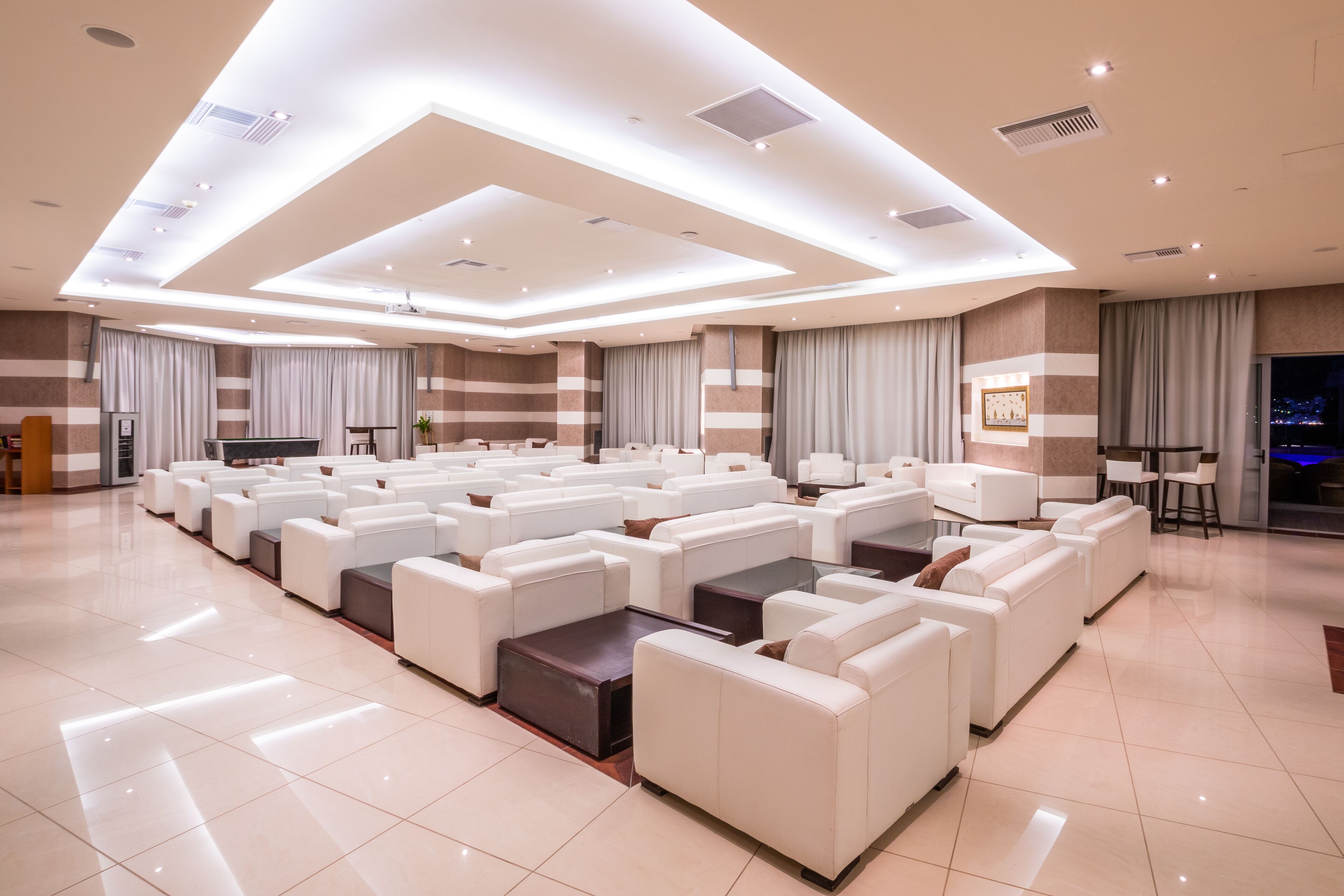 A spacious, well-lit room with multiple white sofas arranged in rows, a large flat-screen TV, and a modern ceiling design.