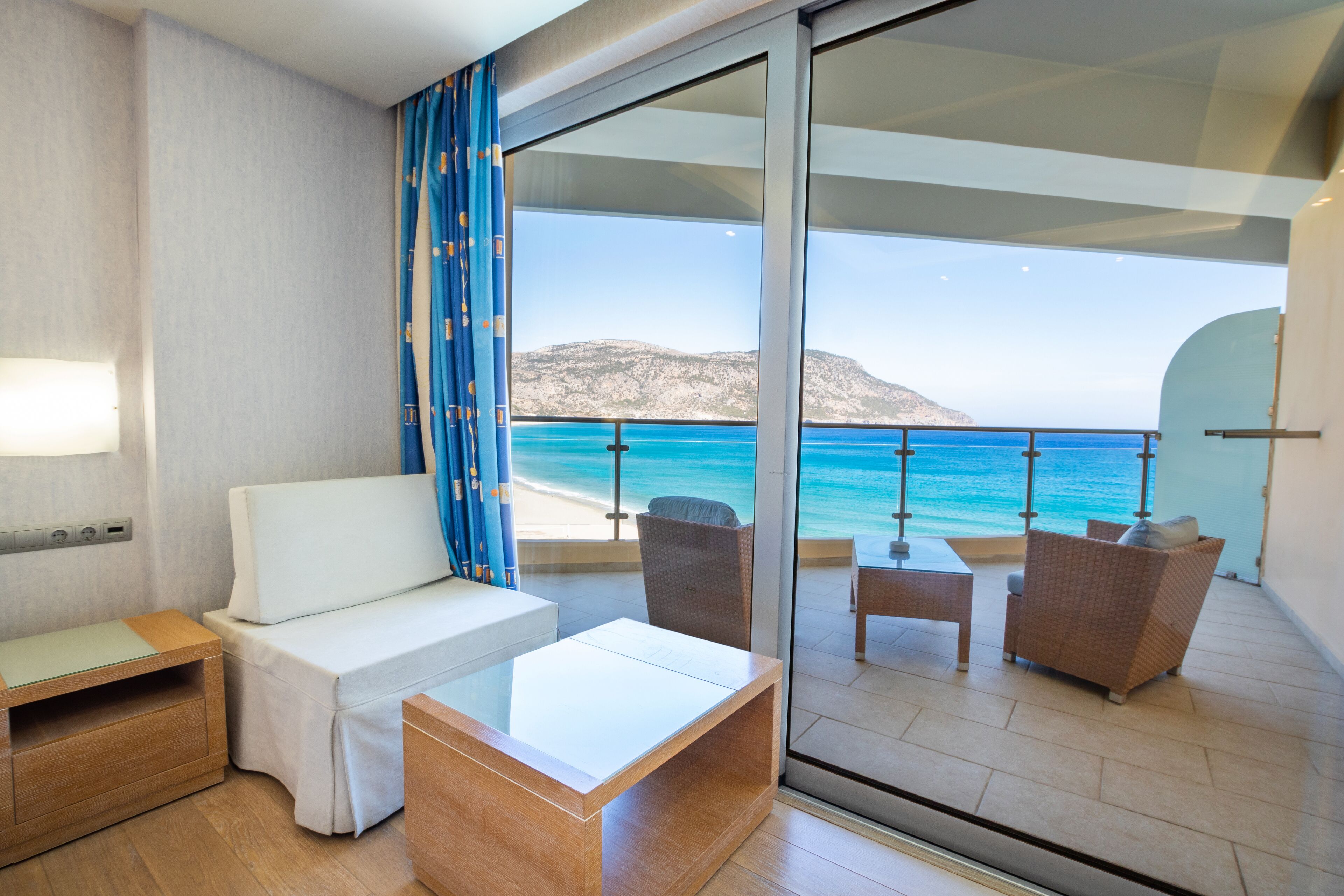 A balcony with a view of the sea and a mountain, furnished with a sofa, chairs, and a table.