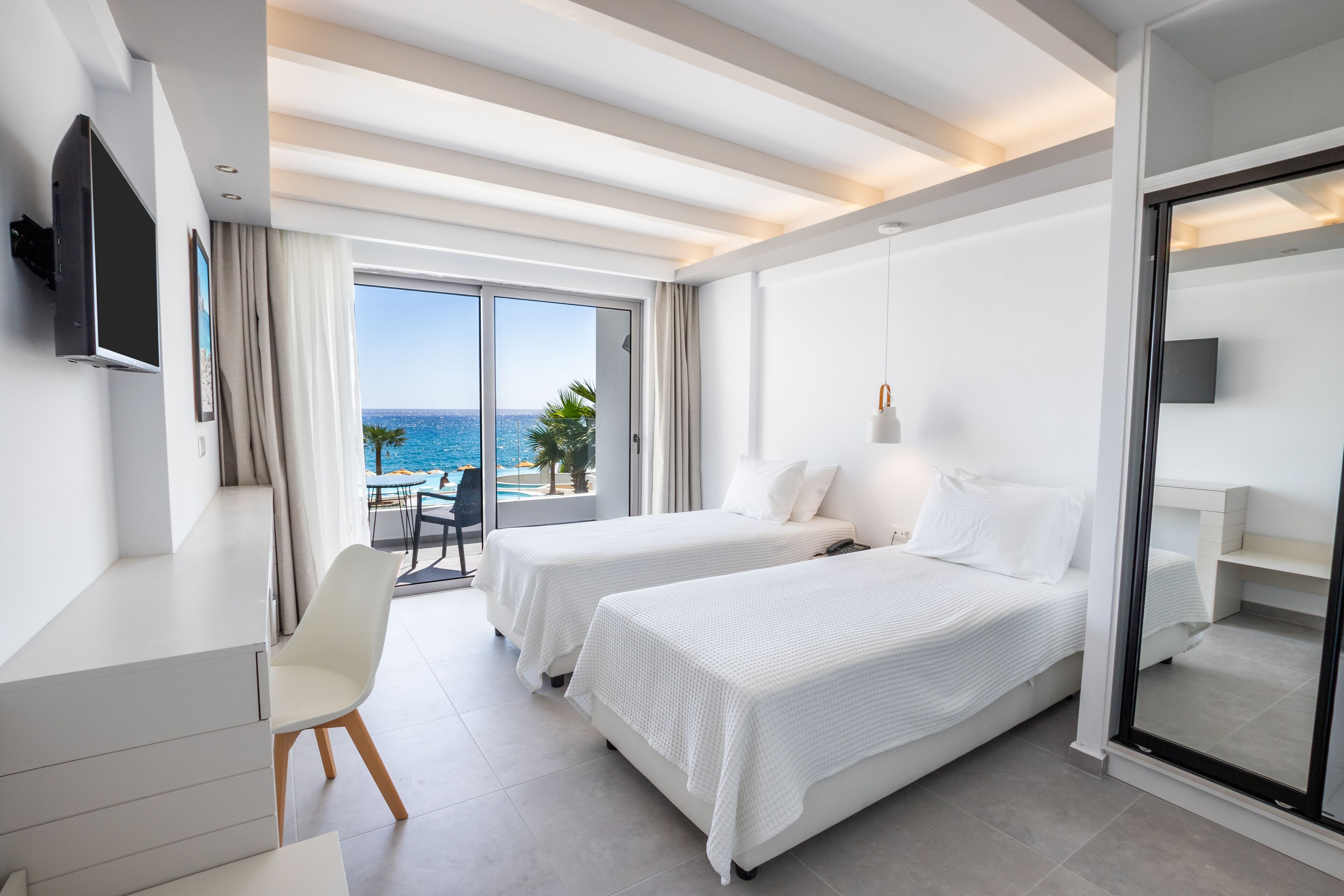 Habitación de hotel moderna con dos camas, un escritorio y una televisión. La habitación tiene un balcón con vista al mar.