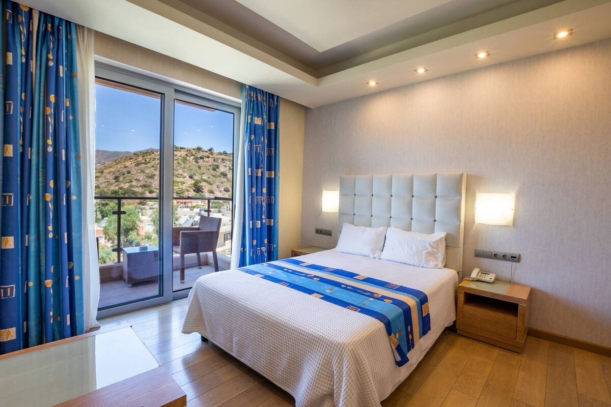A hotel room with a large bed, a nightstand, a desk, and a view of a mountainous landscape through a sliding glass door.