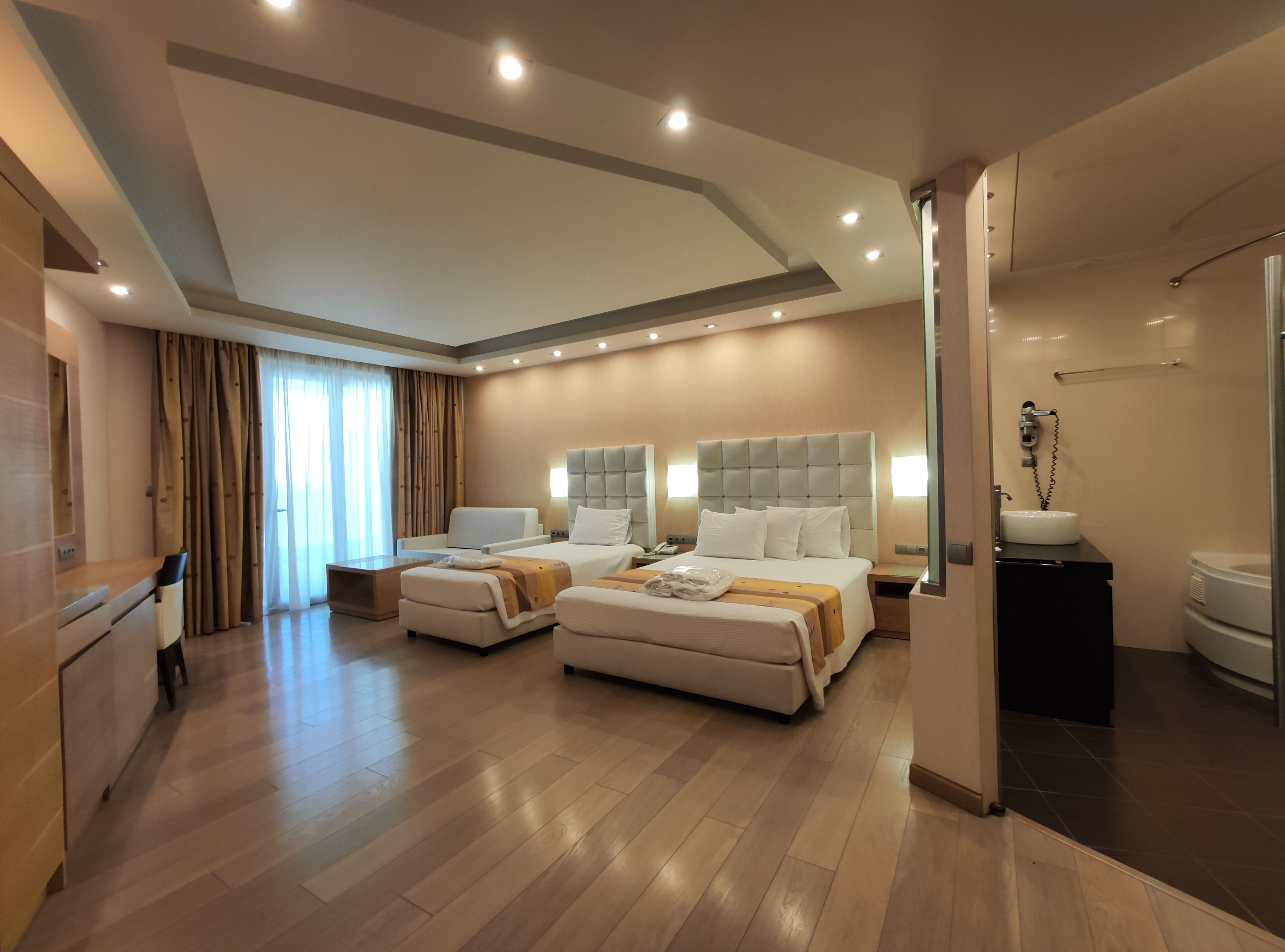 A modern hotel room with two beds, a large window with curtains, a bathroom with a sink and mirror, and a wooden floor.