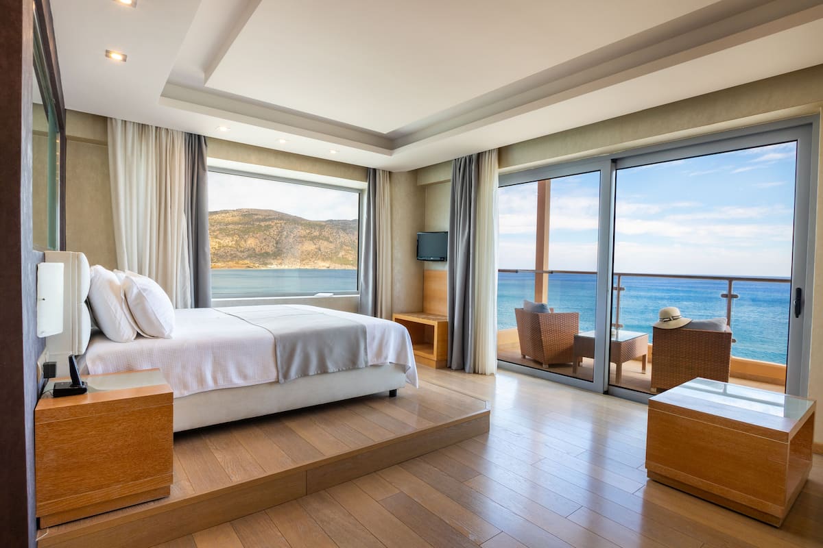 A modern bedroom with a large bed, a TV, and a balcony with a view of the sea.