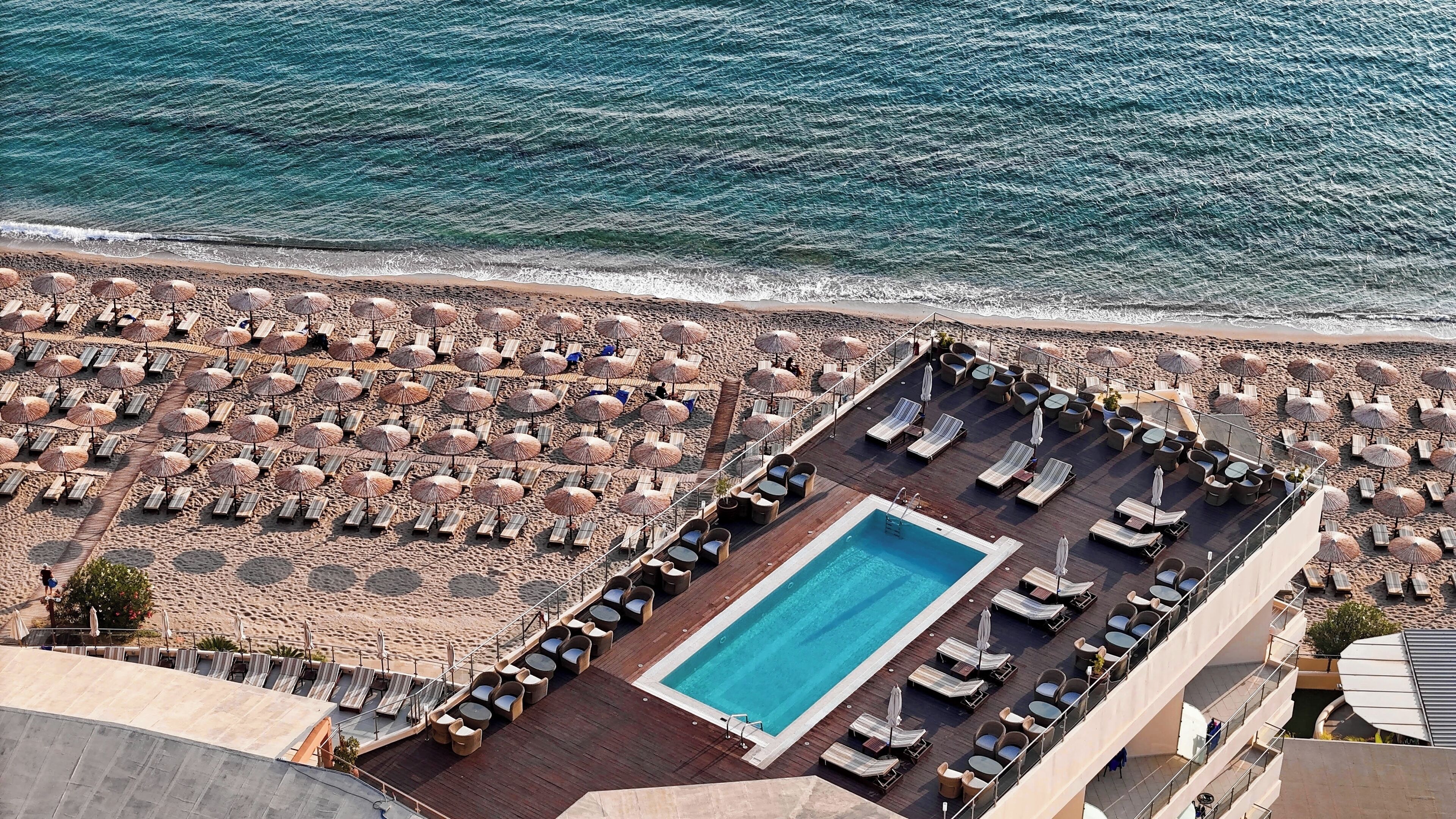 A rooftop pool area with sun loungers and umbrellas, overlooking a beach with umbrellas and lounge chairs.