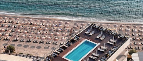 A rooftop pool area with sun loungers and umbrellas, overlooking a beach with umbrellas and lounge chairs.