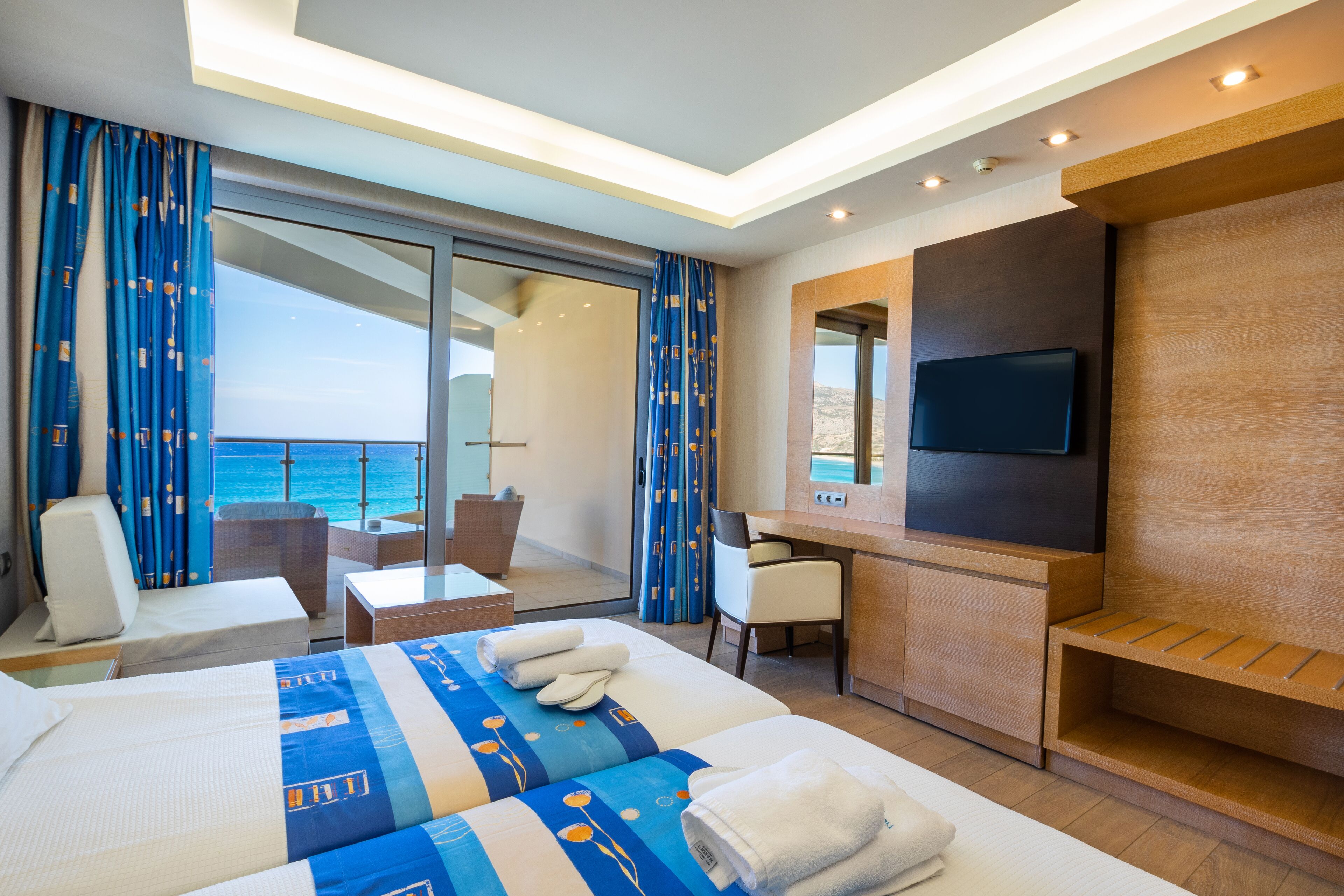 A modern hotel room with a large bed, a desk, a chair, and a TV. There is a balcony with a view of the sea.