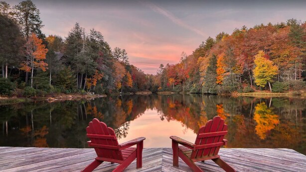 A wooden deck with two red Adirondack chairs facing a calm lake surrounded by autumn-colored trees. - Welcome to A Little Bit O' Heaven! (Lake Toxaway)