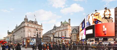 A busy urban square with historic buildings, a prominent red double-decker bus, and a large digital billboard.