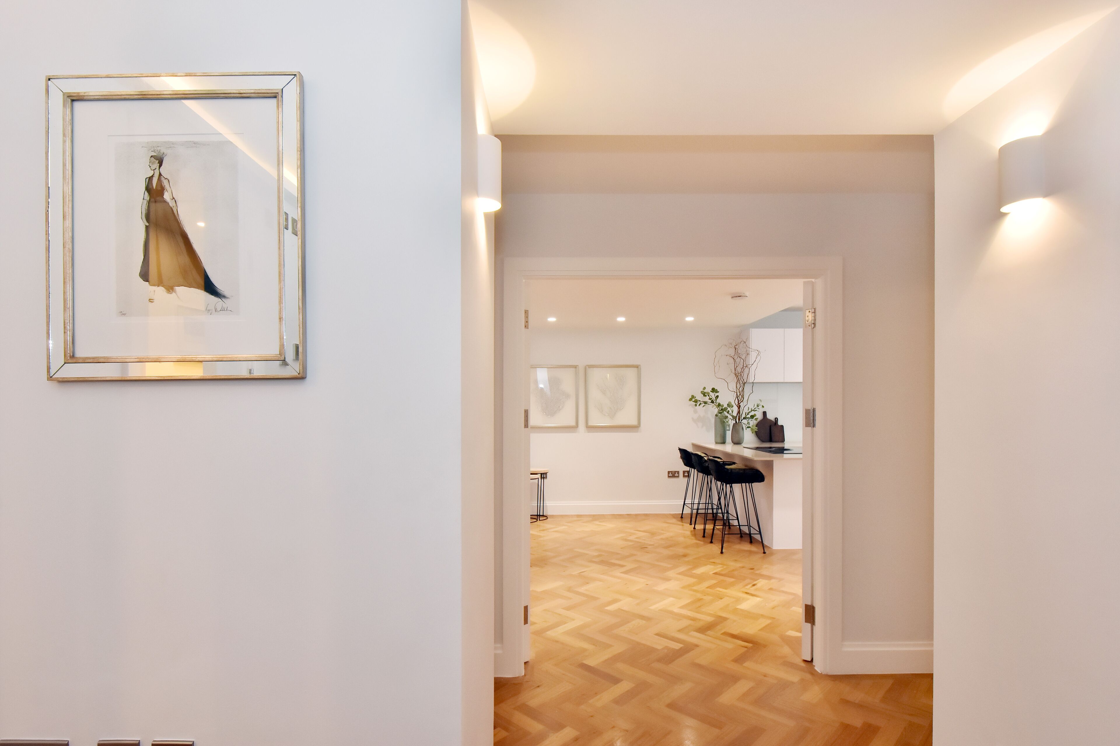 A hallway with a framed artwork, a wooden floor, and a view into a dining area with chairs and a table.