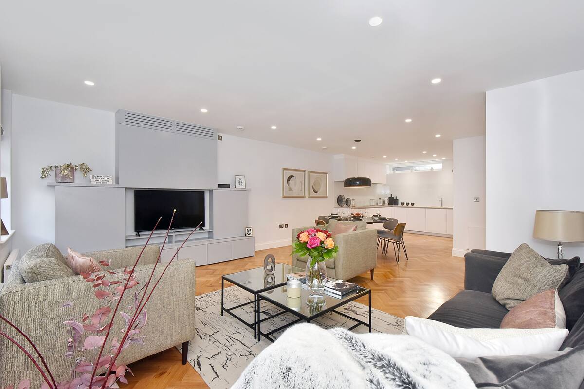 A modern living room with a sofa, coffee table, and decorative plants. A flat-screen TV is mounted on the wall, and a dining area is visible in the background.