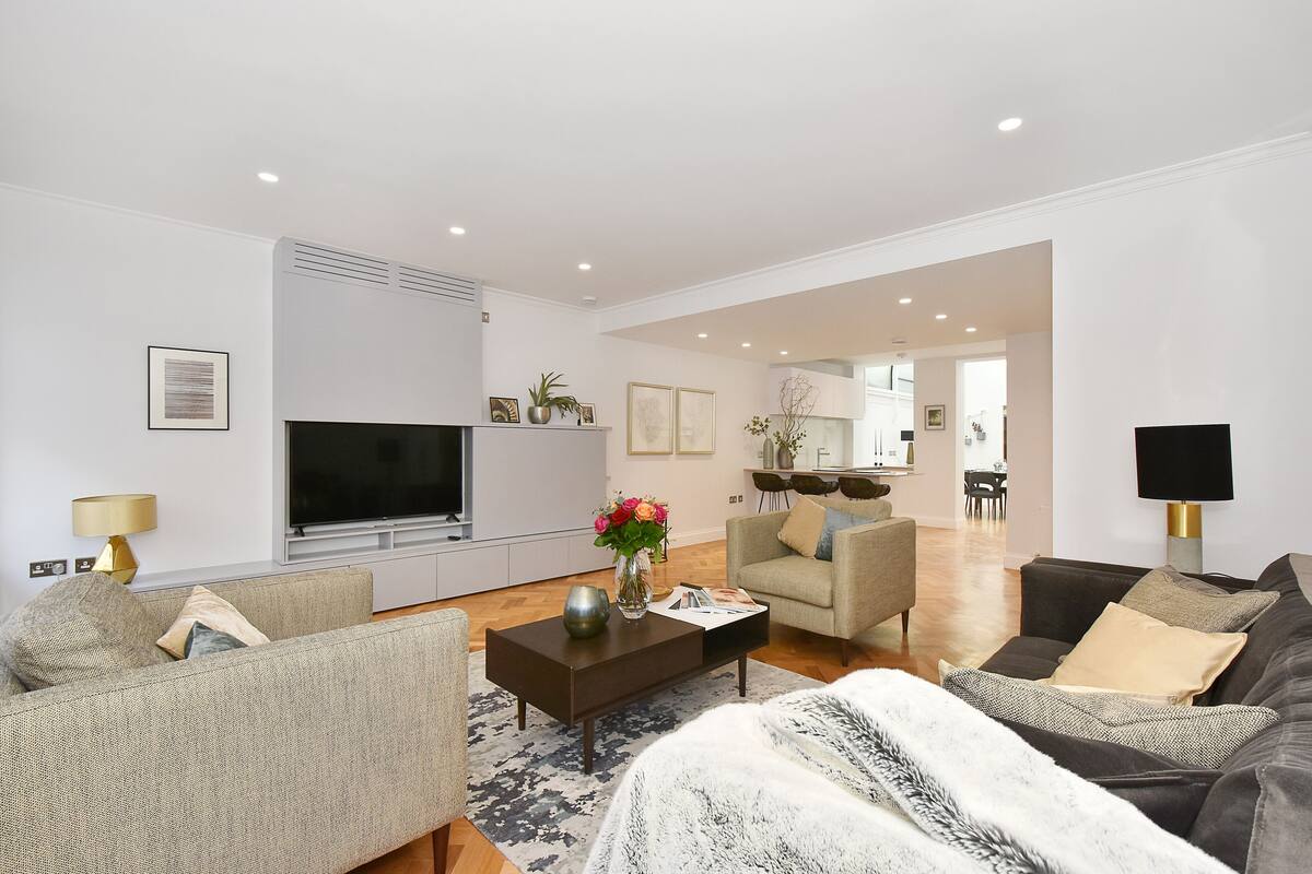 A modern living room with a flat-screen TV, sofas, a coffee table, and a dining area in the background.