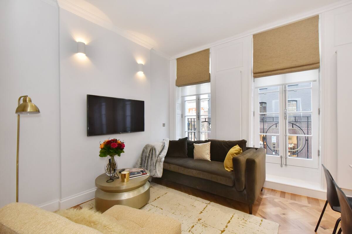 A modern living room with a sofa, a flat-screen TV, a round side table with books and a vase of flowers, and a large window with a view of a balcony.