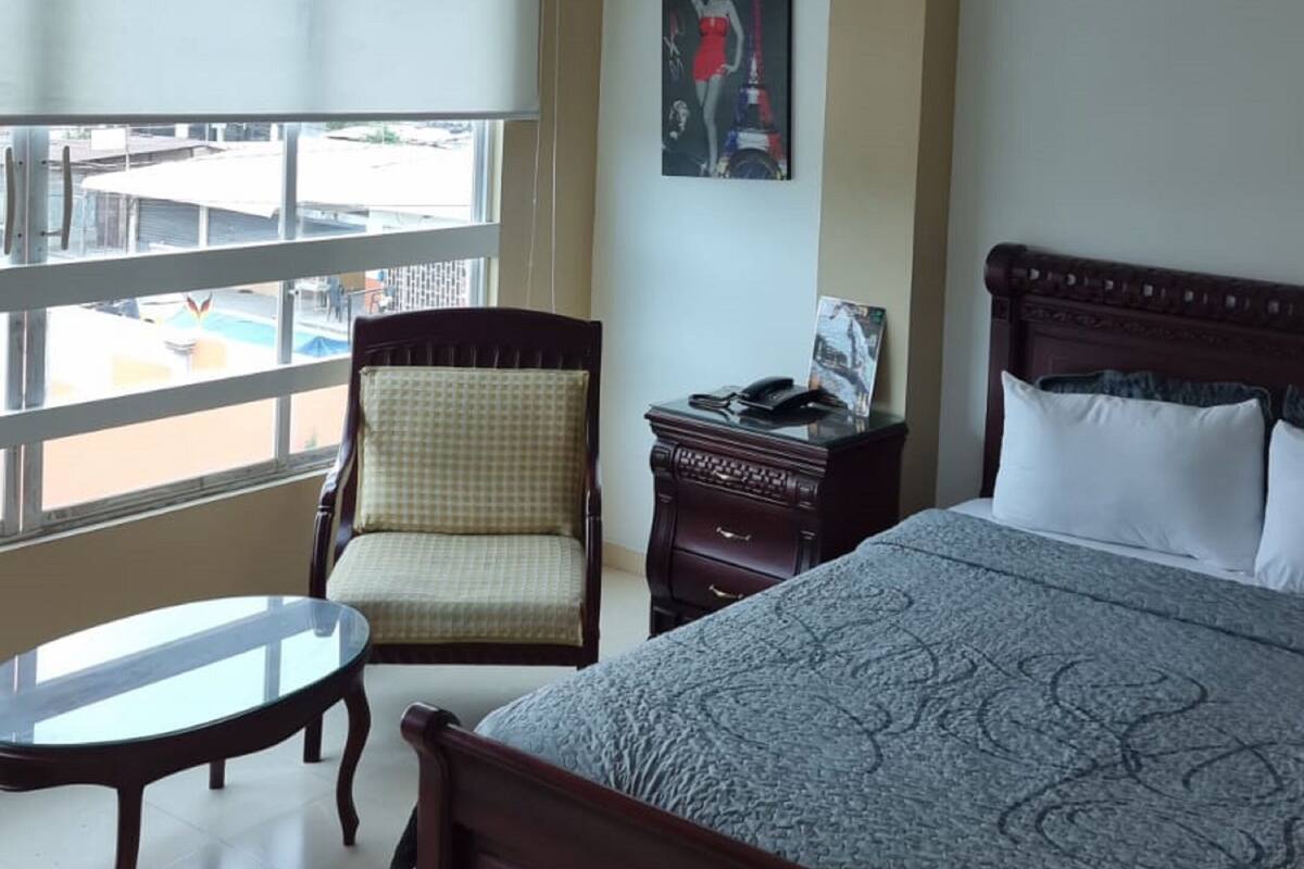 Ein Hotelzimmer mit Bett, Sessel, kleinem Tisch, Nachttisch, einer Chandelieber, einem Fenster mit Blick und einem gerahmten Bild an der Wand.