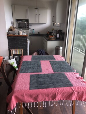 A dining area with a table covered by a pink and black quilt, a chair, and a view of a balcony through a large window.