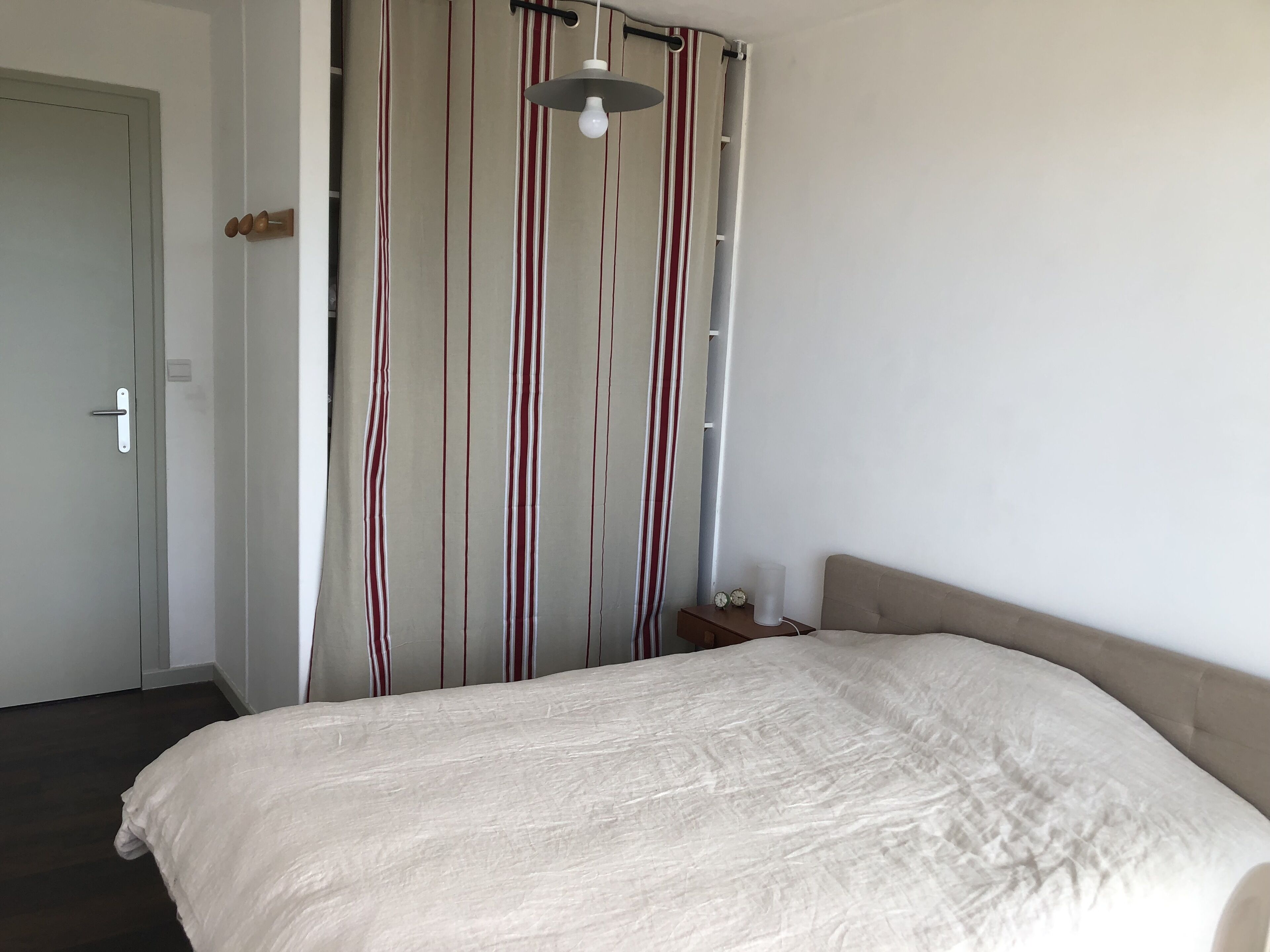 A bedroom with a bed, a curtain with vertical stripes, a bedside table with a lamp, and a door.