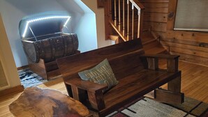 A wooden bench with a cushion, a rustic wooden table, and a patterned rug in a cozy room with wooden flooring and a staircase.