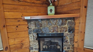 A stone fireplace with a wooden mantel, a bench, and a rug in a log cabin interior.