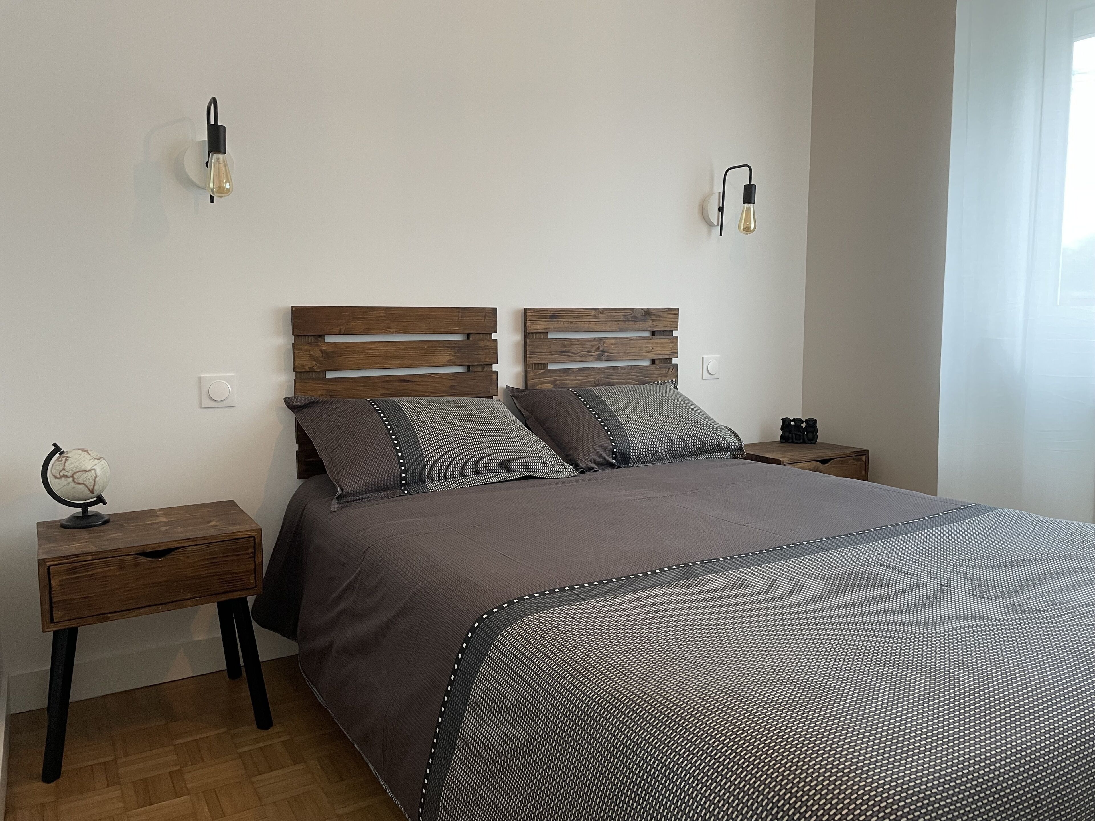 Une chambre à coucher avec une tête de lit en bois, une couvre-lit grise, deux tables de chevet et un globe sur l’une des tables.