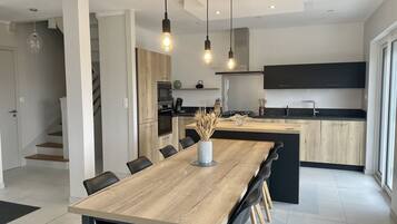Une cuisine moderne avec un îlot central en bois, des armoires noires et un four encastré. On y trouve également un coin repas avec une table en bois et des chaises noires.