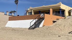 A building with a staircase leading to a balcony, surrounded by sand and palm trees.