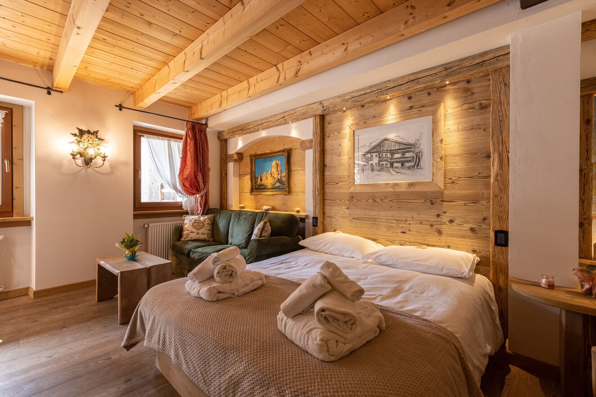 A cozy room with wooden beams, a bed with rolled towels, a green sofa, a small table with a plant, and artwork on the wall.