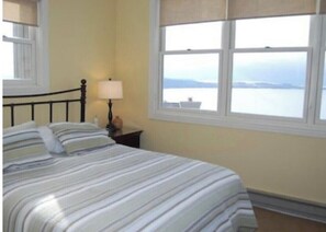 A bedroom with a bed, a nightstand, a lamp, and a window with a view of the sea.