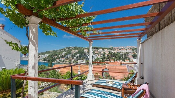 A balcony with a view of a coastal town, a pergola, and lounge chairs.