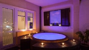 A room with a hot tub, a window with blinds, a wall-mounted shelf with decorative items, and a potted plant.