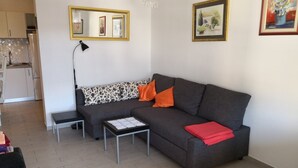 A living room with a grey sofa, a small table with a white doily, and a bench. There is a mirror on the wall and a painting above the sofa.
