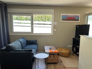 A living room with a sofa, a small table, a wooden chest, a television, and a window with a view of greenery.